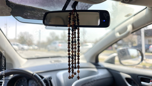 Car interior with hanging wooden beads on rearview mirror. Beads dangle from mirror's mounting hook. Driver's side dashboard and steering wheel visible. Sunlight filters through windshield