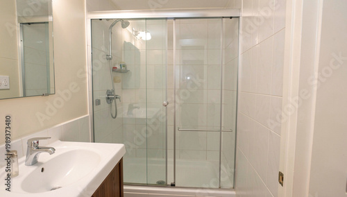 Modern bathroom with glass shower enclosure and sink. Clean white tiles line the walls and floor. Showerhead and faucet are visible above the sink. Glass doors frame the shower area