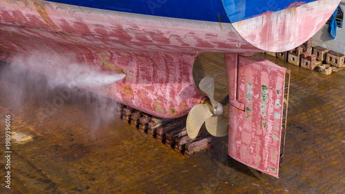 massive cargo ship hull, propeller, and rudder in a dry dock during maintenance. High-pressure water jet cleaning the red surface. Marine engineering, shipyard repair, heavy industry.