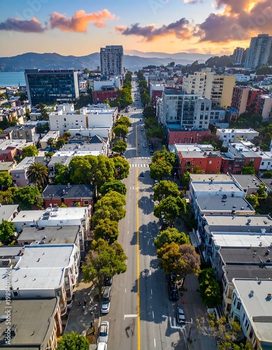 Aerial View Of A Long City Street At Sunset With Dense Urban Architecture Green Trees And A Distant Bay Under A Colorful In A Coastal City