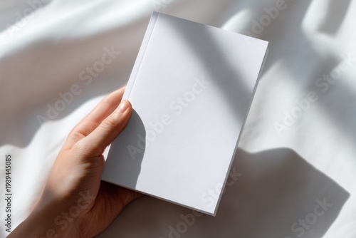 A person's hand holding a blank white book cover. Minimalist mockup template for design and branding on a soft fabric background