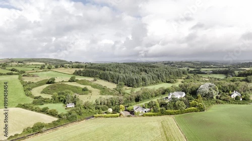 A picturesque rural scene unfolds, showcasing agricultural land, a dense forest, and scattered dwellings under a cloudy sky.