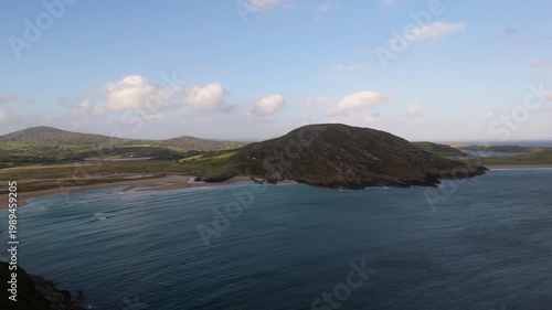 Soaring over the rugged Irish coastline showcasing green mountains, a serene bay, and sandy beaches under a bright sky.