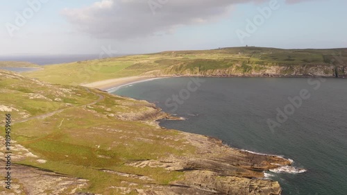 Expansive view captures a beautiful sandy beach nestled between rugged coastal cliffs and rolling green hills. Calm ocean waters reflect the soft light of a sunny late afternoon sky.
