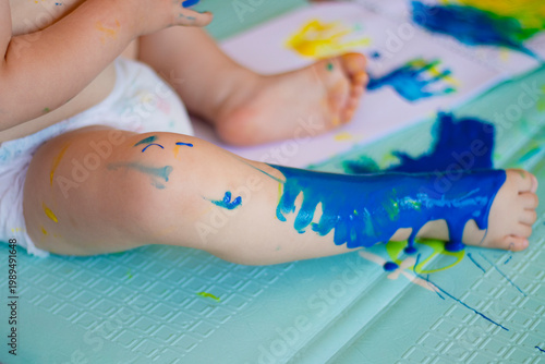 Infant explores with vibrant paints. Cute baby indulges in paint play activity. Chubby baby seated on textured mat with colorful paint splatters and finger marks