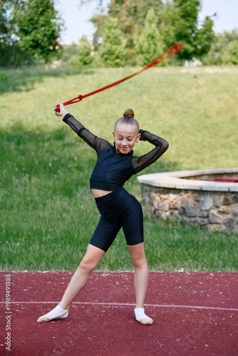Young girl performs rhythmic gymnastics with a ribbon on a sports field outdoors, showcasing athletic skills. Young Girl Practicing Rhythmic Gymnastics Outdoors on Sunny Day