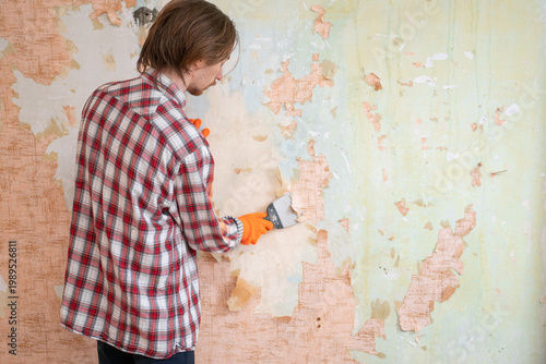 Man in plaid shirt removing old wallpaper from wall preparing room for apartment renovation process