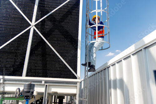 A male engineer with safety vest and helmet climbs a ladder to inspect machine of rooftop cooling tower. A male technician worker climbing a ladder to check and verify the building cooling system.