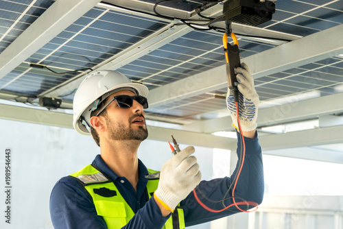 A male engineer with safety vest and helmet works with electrical instruments on photovoltaic solar panels. Technician uses multimeters to check performance of rooftop solar power system at solar farm