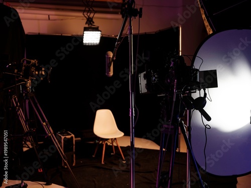 Warm lights illuminate a sleek podcast studio filled with cameras and microphones, creating an inviting space for engaging interviews. The empty white chair awaits its guest, inspiring anticipation