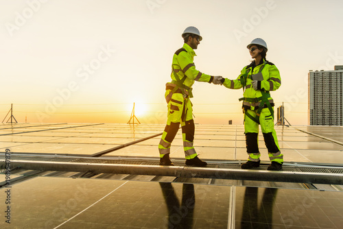 Engineers in safety clothing with helmet have handshake during work at solar farm rooftop. Man and woman workers shake hands with city view at sunset after finishing installation solar panels on roof.