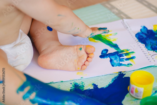 Baby paints on mat, tiny hands exploring blue green paint, colorful handprints and footprints, small paint pot tipped, playful messy texture, soft natural