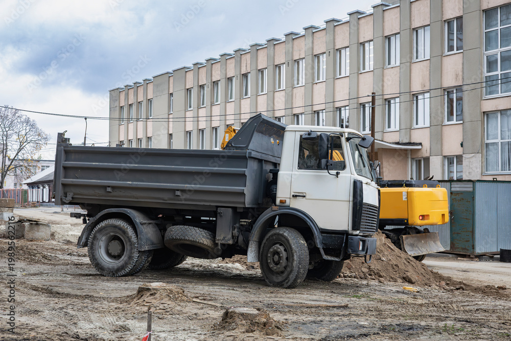 Obraz premium A dump truck is parked on a construction site surrounded by machinery, dirt piles, and a large building under overcast skies. Workers are present in the area