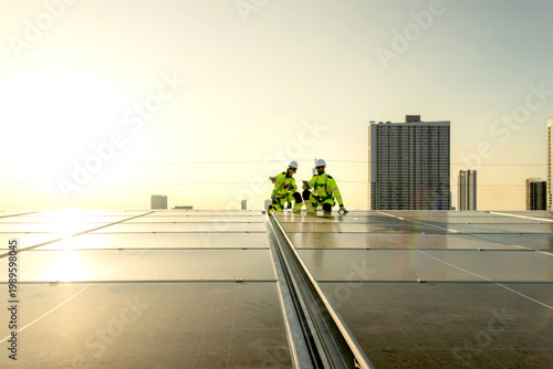 Two engineers in safety clothing with helmet check solar system at rooftop solar farm with city view. Engineers installing solar panels. Man and woman workers inspecting solar panels on roof at sunset