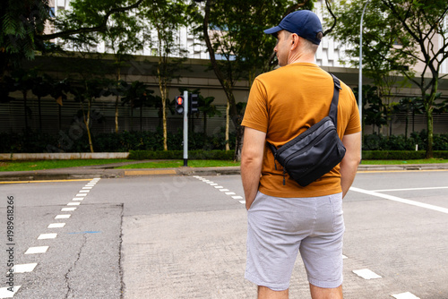 Man from behind wearing casual clothes and black sling bag stands at urban street crossing. Pedestrian waiting at red traffic light in city environment.