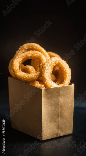 Golden, crispy onion rings stacked in a kraft paper box against a dark background.
