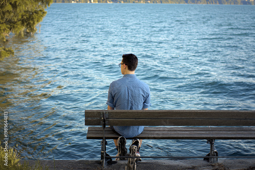 A young man gazes thoughtfully at the water from a bench, surrounded by nature's serene beauty.