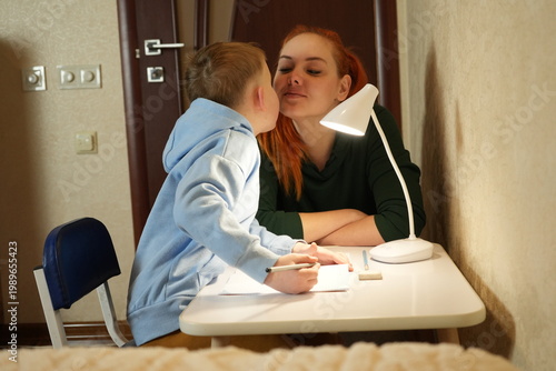Mother and child sharing a tender moment and kissing while doing homework together. Family love, parenting, support and home education concept.