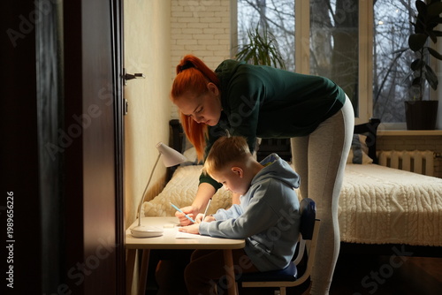 Mother leaning over her son and helping with homework at a desk under warm lamp light. Cozy home atmosphere, parenting, education and family support concept.