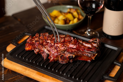 Sliced beef tagliata with tongs on cast iron grill pan with roasted potatoes and red wine