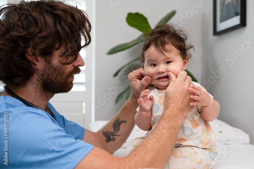 Canvastavla Male pediatrician in blue scrubs gently checking a small child's mouth and jaw