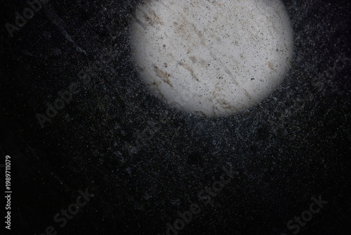 Moon glowing behind the details of a dirty window. White, textured circular object against dark background. Dust particles float around. Abstract, close-up shot with soft lighting. Natural, organic su