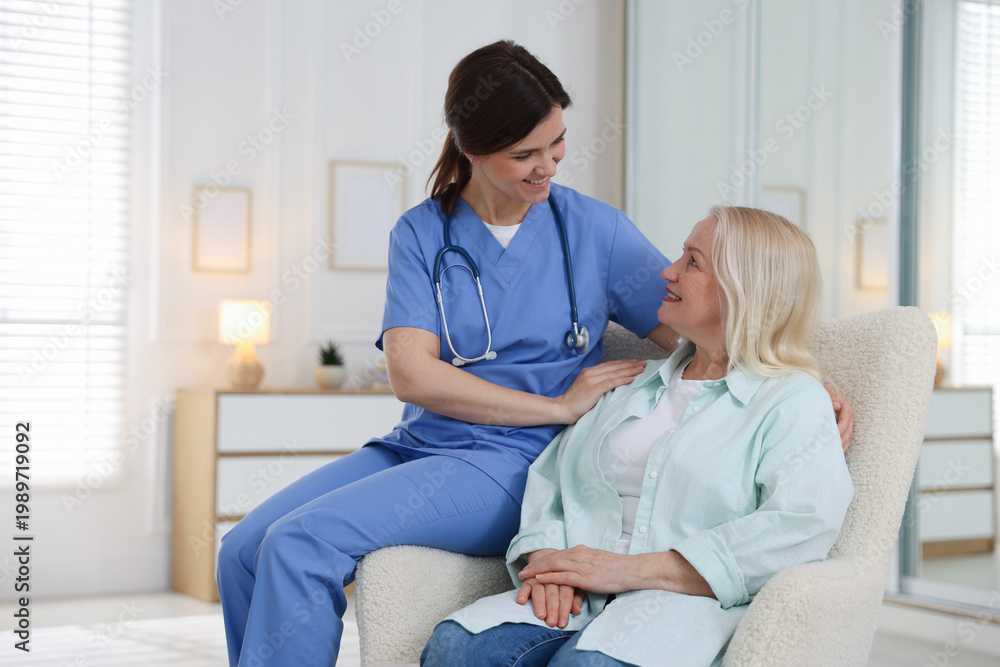 Fototapeta premium Smiling care worker with senior woman on armchair at nursing home. Space for text