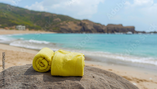 Two light yellow towels on a gray rock by a turquoise sea with gentle waves, sandy shore, and blurred rocky hills under a partly cloudy sky—calm, scenic summer beach atmosphere