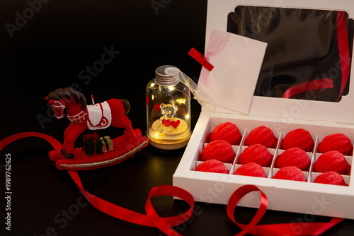 A white cardboard box with red shortcrust pastry nuts, with filling, on a black background, decorated with red ribbons and toys - a horse and a teddy bear in a jar