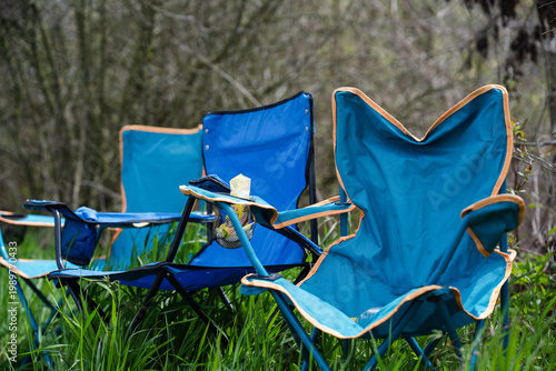 Billede på lærred A row of blue foldable camping chairs with cup holders set up on a lush green lawn