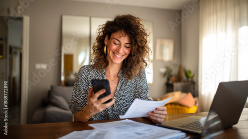 Woman sits at home table checking bills on smartphone while smiling during daytime