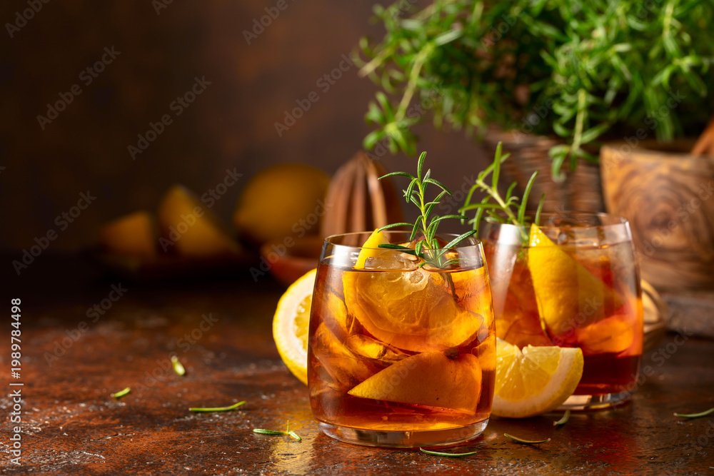 Fototapeta premium Iced tea or cocktail with ice, rosemary and lemon slices on the old brown table.