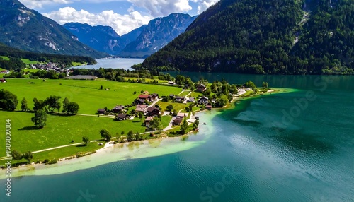 Aerial view of a lakeside village nestled between green fields, turquoise water, and towering, lush green mountains under a blue sky