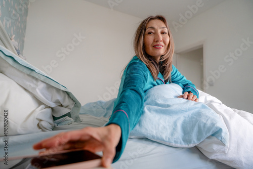 Fotografi Woman reaching for her phone while sitting on a bed in a bright room during morn