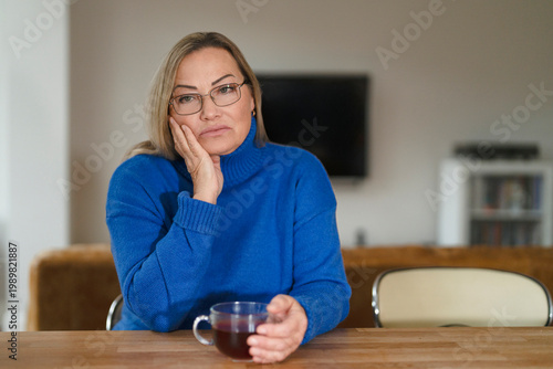 Fotografie Woman sitting at kitchen table holding cup of tea and looking thoughtful in mode