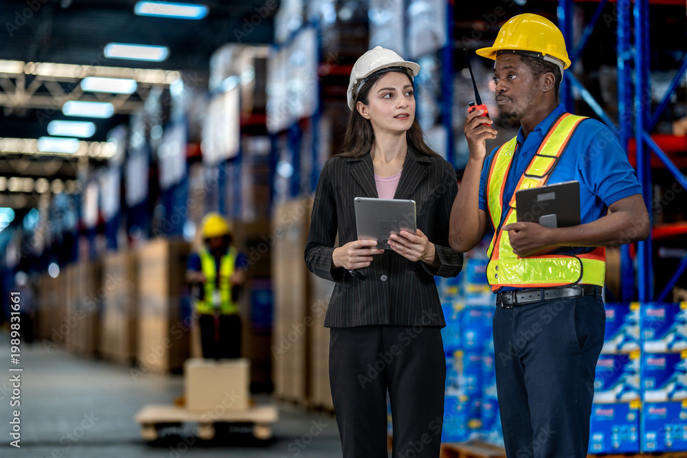 Fototapeta premium Diverse warehouse team collaborating in a large distribution center. A worker operating a pallet jack while a manager and colleague inspect digital inventory records together.