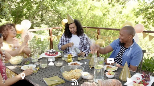 Grabbing bottle, center guest popping cork, then adjusting gold balloon while friends celebrating