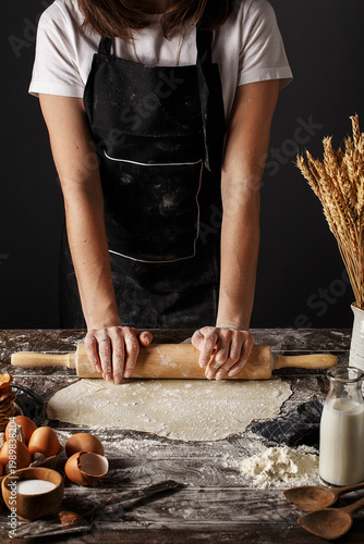 Konstfotografi Woman in black apron rolling out dough on wooden table with spilled flour, raw eggs, bottle of milk and wheat spikes