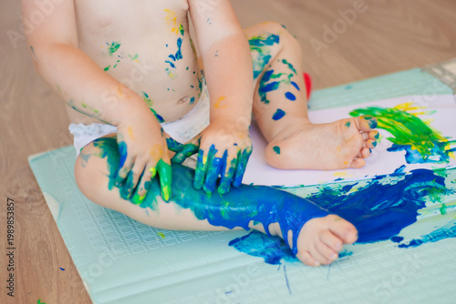 Baby paints on mat, tiny hands exploring blue green paint, colorful handprints and footprints, small paint pot tipped, playful messy texture, soft natural