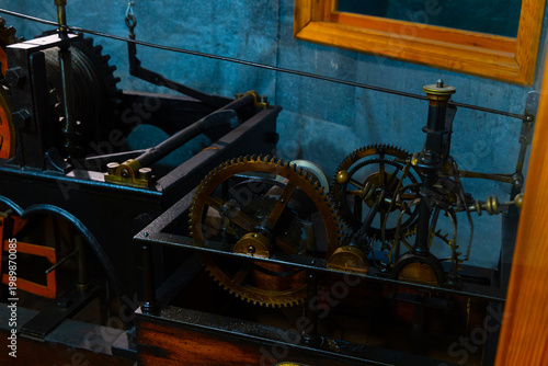 Intricate mechanical device with brass and black metal gears on display against a blue wall with a wooden frame