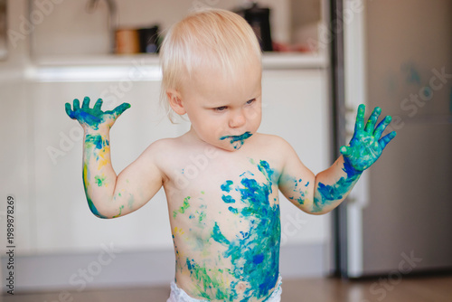 Baby covered in blue green paint, exploring colorful mess with hands raised, joyful expression, diapered toddler in bright kitchen, natural light, playful