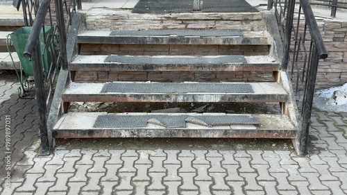 Worn antiskid coating on steps at pickup entrance, rusted metal treads with peeling paint and grime, uneven concrete pavers, damp.