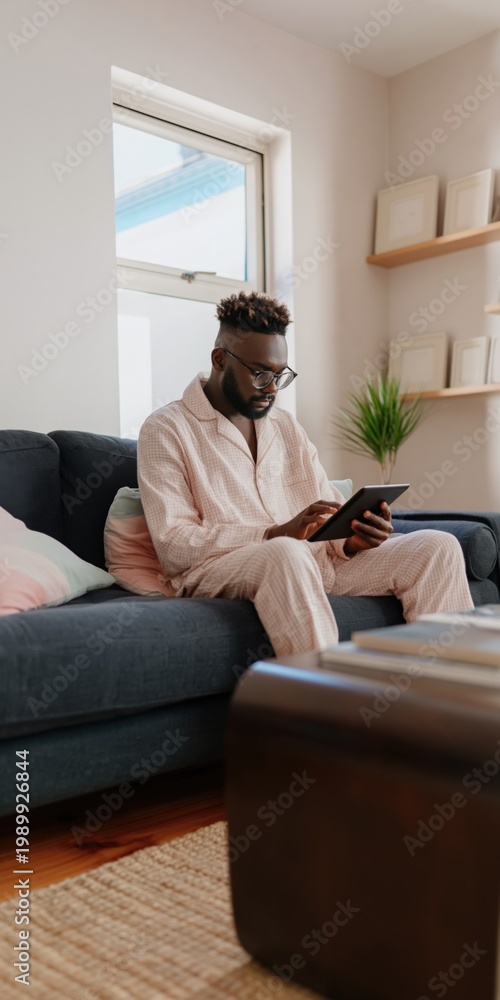 Obraz premium Black man using tablet seated on living room sofa near window