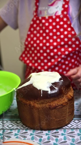 Close-up of a child in a red polka-dot apron carefully spreading thick white icing over a freshly baked dark brown Easter kulich using a spoon. Sweet and joyful moment of decorating traditional Easter