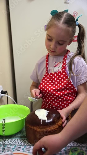 Cute young girl in a red polka-dot apron carefully spreads white icing on a freshly baked Easter kulich. She is focused and enjoying the process. Bright green bowl with icing on the table. 