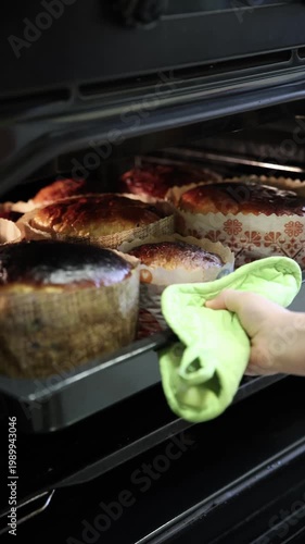 Hand wearing a bright green oven mitt carefully removes a tray of golden-brown freshly baked Easter kulich paska from the hot oven. Traditional Ukrainian and Eastern European Easter bread with a beaut
