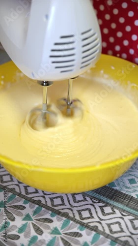 Close-up of a white electric hand mixer with spinning beaters mixing thick pale yellow batter inside a bright yellow mixing bowl. Swirls and smooth texture visible. Home baking process on a patterned 