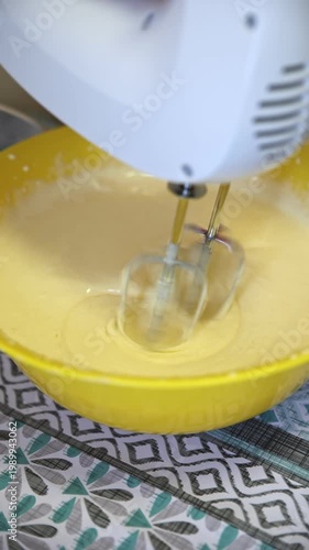 Close-up of a white electric hand mixer with spinning beaters mixing thick pale yellow batter inside a bright yellow mixing bowl. Swirls and smooth texture visible. Home baking process on a patterned 