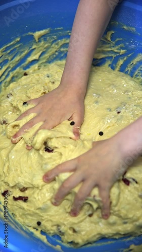 Close-up of a children hands deeply immersed in thick yellow dough with raisins inside a large bright blue bowl. Sensory baking activity, messy and joyful moment of kids helping in the kitchen.