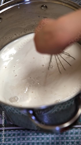 Close-up of a child’s hand holding a metal whisk and actively stirring a thick white liquid inside a large stainless steel pot. Bubbles and creamy texture visible. Cozy home cooking scene on a pattern